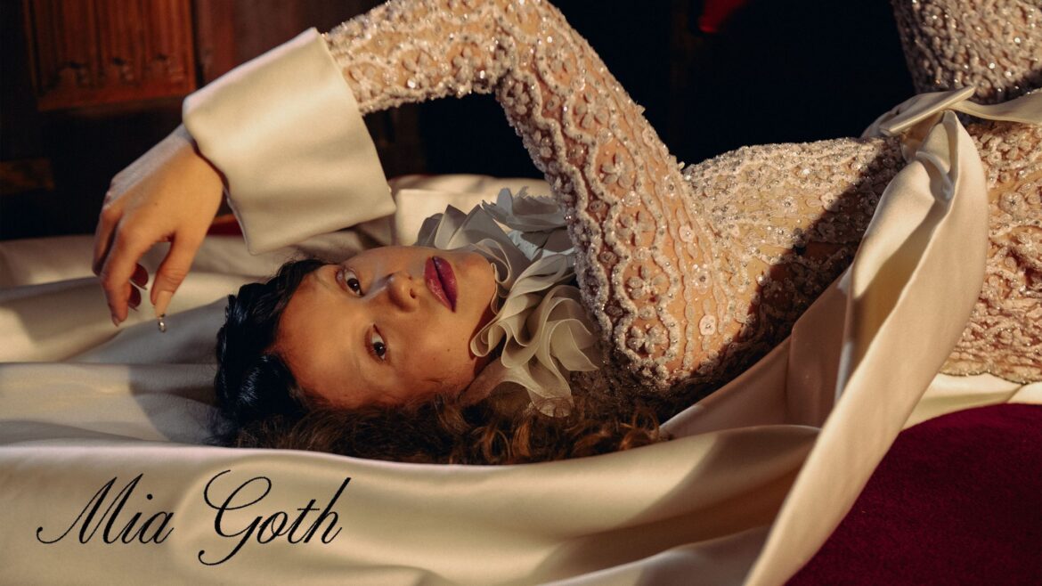 Hero image for the October cover story with Mia Goth. She is laying on the ground looking to the camera wearing a white crystal embellished bodysuit with ruffled collar detailing. 